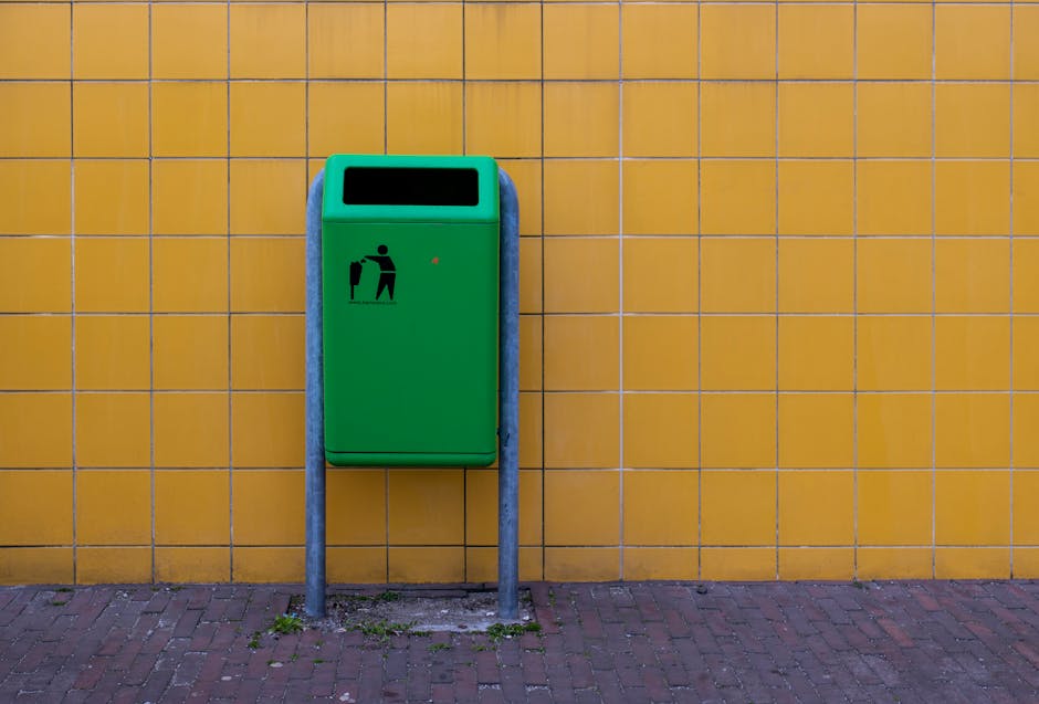 A green rubbish bin with a black pictogram of a person discarding waste is mounted on a yellow tiled wall outside a building. The bin is supported by two silver metal posts fixed into a paved red brick ground. There is a small patch of grass and dirt around the base of the posts. The bin's lid is closed, and a small red indicator light or tag is visible on its front. The scene is illuminated by natural light, suggesting daytime. This image relates to home relocation or moving logistics, as part of furniture disposal or recycling process during house removals, with [COMPANY_NAME] providing related services for furniture transport and recycling in Pimlico.