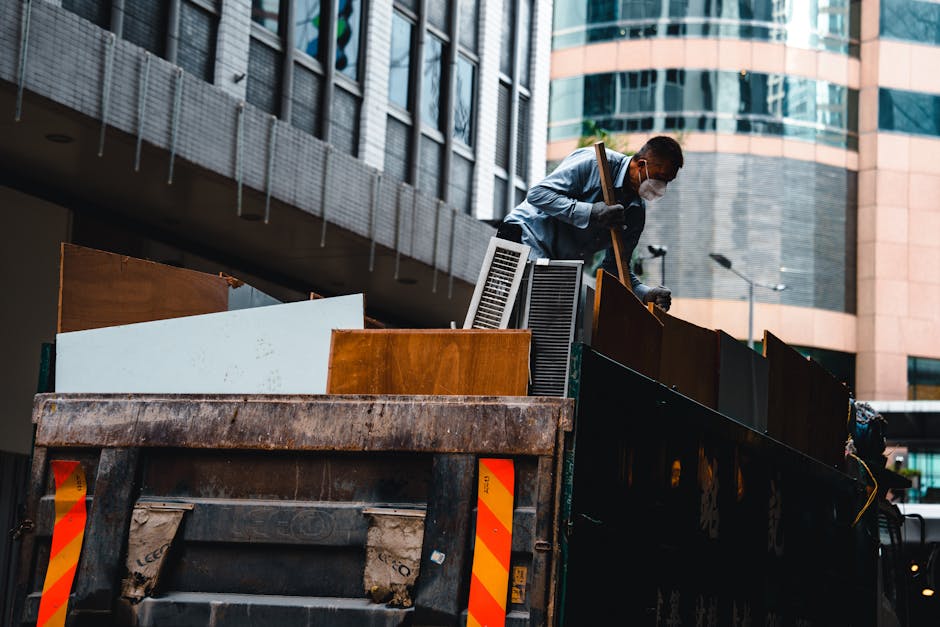 A worker wearing a face mask and gloves is standing on the back of a large, open-top truck loaded with bulky furniture and household items. The truck is situated outdoors on a city street, with modern multi-storey buildings featuring glass and concrete facades visible in the background. The furniture includes wooden cabinets, screens, and large objects covered in cardboard or wrapped in protective materials. The worker appears to be in the process of loading or unloading the furniture, using a wooden stick or lifting tool. The truck has a worn appearance with visible rust and dirt on its metal surface, and safety markings are visible on the rear. The scene captures a home relocation or furniture transport operation in an urban environment, aligning with professional removals and recycling services provided by Man and Van Pimlico.