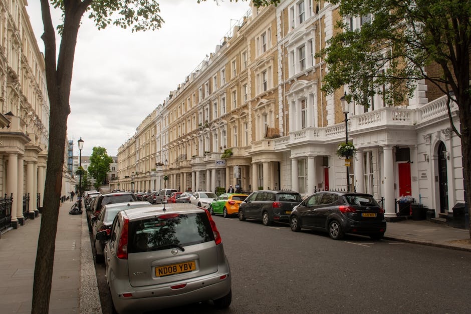 A street view on Eccleston Street in Pimlico showing a row of white and beige Victorian-style terraced houses with decorative facades and bay windows. Several cars, including a grey hatchback with the registration ND08 YBV, are parked parallel along the curbside on both sides of the street. There are trees with green foliage lining the pavement, and a lamppost near the center of the image. The sky is overcast, casting diffused light over the scene. The image captures the residential street during the daytime, with no visible people or moving vehicles, illustrating typical Victorian home architecture and urban parking in Pimlico. This setting aligns with house removals and furniture transport services provided by Man and Van Pimlico, highlighting the environment where home relocation and packing processes take place.