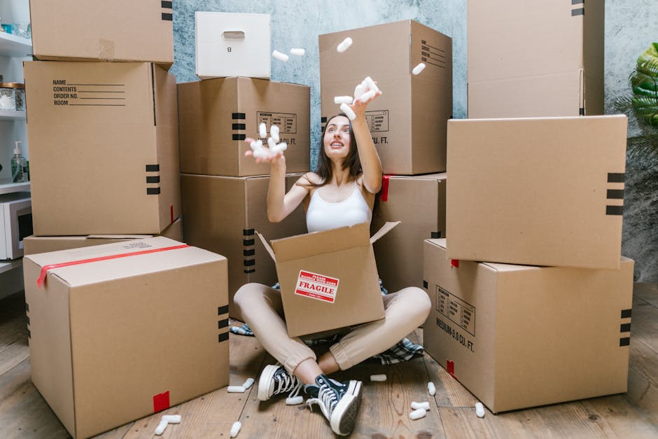 A woman sitting cross-legged on a wooden floor surrounded by large cardboard moving boxes, some with red and black labels, and open one revealing packing material. She is wearing a white tank top, beige trousers, and striped sneakers, with long dark hair. She is smiling and tossing packing foam into the air, indicating unpacking or packing activities. The boxes are stacked against a textured grey wall, and a small portion of a kitchen or office area with shelves and a plant is visible in the background. This scene depicts house relocation and packing processes, with practical moving equipment like boxes and packing materials, typical of services offered by Man and Van Pimlico, supporting efficient furniture transport and home removals.