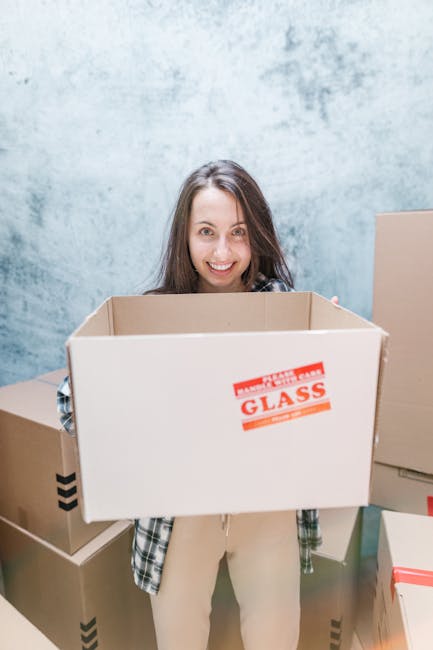 A woman with long dark hair smiling and holding an empty cardboard box labeled 'GLASS' on the side, standing indoors against a light blue textured wall. Around her are several other cardboard boxes, some sealed with red and black tape, and one partially visible behind her. The scene appears to be part of a house removal or packing process, with the woman preparing for or engaged in moving furniture and belongings. The environment is well-lit, and the boxes are made of brown corrugated cardboard, with some wrapped in plastic for protection, typical in professional relocation services like Man and Van Pimlico involved in rapid home relocation and furniture transport.