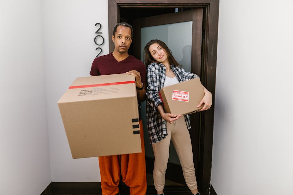 A man and a woman standing inside a doorway, holding cardboard boxes used for home relocation; the man is carrying a large, plain cardboard box with a shipping label, dressed in orange pants and a maroon shirt, while the woman holds a smaller box with 'Fragile' and handling labels, wearing beige trousers and a plaid shirt; both are positioned in a narrow corridor with a plain white wall on one side and a dark wooden door frame behind them, indicative of a residential setting during a furniture transport or packing and moving process; the scene is well-lit, capturing the typical loading and packing activities associated with professional removals, with visible attention to safety and organisation, reflecting the services provided by Man and Van Pimlico in rapid house removals and logistics.