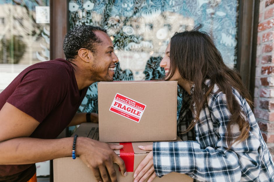 A woman with long dark hair smiling and holding an empty cardboard box labeled 'GLASS' on the side, standing indoors against a light blue textured wall. Around her are several other cardboard boxes, some sealed with red and black tape, and one partially visible behind her. The scene appears to be part of a house removal or packing process, with the woman preparing for or engaged in moving furniture and belongings. The environment is well-lit, and the boxes are made of brown corrugated cardboard, with some wrapped in plastic for protection, typical in professional relocation services like Man and Van Pimlico involved in rapid home relocation and furniture transport.
