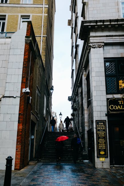 A narrow cobbled street in Pimlico with ascending stone steps between tall, closely spaced buildings of mixed brick and stone facades. On the steps, there are several pedestrians dressed in dark clothing, some carrying bags or belongings, and a red umbrella resting on the lower part of the staircase. To the right, a building features a sign for 'The Coal Hole' pub, with decorative elements and illuminated signage, while the left side shows a building with a white and red brick exterior, incorporating security cameras. The scene is set during daylight with overcast skies, capturing the confined access typical of historic London streets, relevant to house removals and furniture transport in tight urban environments, as managed by Man and Van Pimlico.