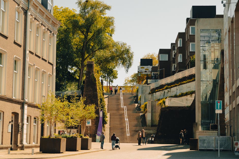 A city street scene showing a series of outdoor concrete stairs leading up a slope, with a metal handrail running along the center. At the base of the stairs, there is a paved pedestrian area with several people walking, some pushing strollers or carrying bags. On the left side, a beige brick building with multiple white-framed windows and a small tree planted nearby is visible. On the right, modern glass-fronted buildings with concrete and metal facades are present, with planters containing yellow flowers placed along the steps' edges. Trees with green foliage line the top of the stairs, and a clear sky suggests daytime. The scene is well-lit with natural sunlight, capturing a typical urban environment suitable for local home relocation and furniture transport activities, as carried out by [COMPANY_NAME], supporting efficient packing and loading processes in tight residential street access conditions.