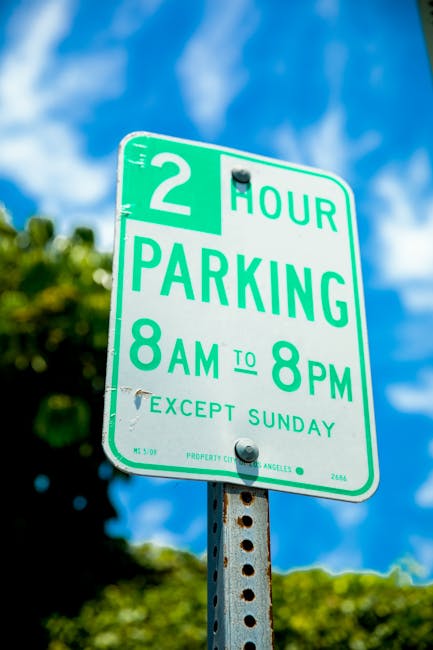 A green and white parking restriction sign mounted on a rusted metal post outdoors, displaying a two-hour parking limit from 8 am to 8 pm, with additional text indicating exceptions on Sundays. The sign is photographed against a bright blue sky with some clouds and blurred green foliage in the background. This type of sign is commonly seen on city streets where parking restrictions are regulated, and it is relevant for home relocation and moving services, such as those offered by Man and Van Pimlico, to ensure proper parking permits and adherence to council rules during furniture transport and packing and moving activities.
