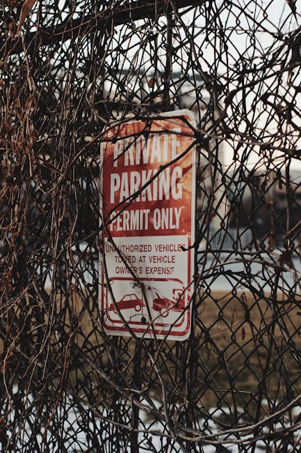 A close-up image of a red and white parking permit sign attached to a metal fence that is overgrown with dry, tangled brown vines. The sign reads 'Private Parking Permit Only' with additional text indicating restrictions against unauthorized vehicles at vehicle owners’ expense. The background appears to show a parking area or street at dusk with muted lighting. The scene suggests a restricted parking zone typically associated with house removals and relocation logistics, with the sign indicating parking control measures typically relevant to moving services, such as [COMPANY_NAME], that may require permits or to manage parking for moving vans. The overall setting emphasizes the importance of following local council rules for parking permits during home relocations in the Pimlico area, aligning with the page about 'Pimlico removal permits & parking suspensions' at [PAGE_URL]. The image demonstrates the environment and logistical considerations involved in home moving and furniture transport in urban settings.