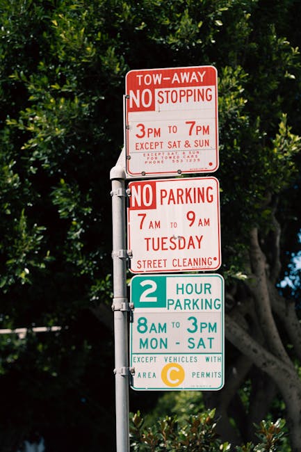 A green and white parking restriction sign mounted on a rusted metal post outdoors, displaying a two-hour parking limit from 8 am to 8 pm, with additional text indicating exceptions on Sundays. The sign is photographed against a bright blue sky with some clouds and blurred green foliage in the background. This type of sign is commonly seen on city streets where parking restrictions are regulated, and it is relevant for home relocation and moving services, such as those offered by Man and Van Pimlico, to ensure proper parking permits and adherence to council rules during furniture transport and packing and moving activities.