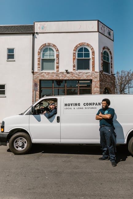 A white moving van parked in front of a two-storey building with brick and white exterior walls, featuring three arched windows on the upper floor. Inside the van, a person wearing a beanie and jacket