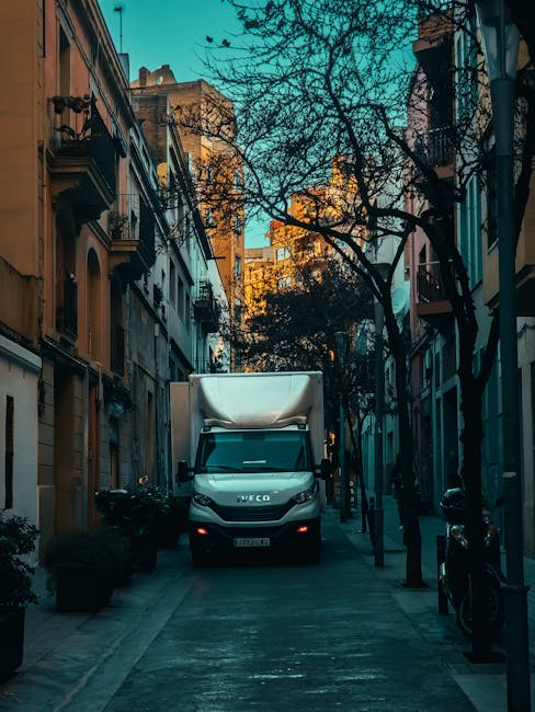 A white moving van parked in front of a two-storey building with brick and white exterior walls, featuring three arched windows on the upper floor. Inside the van, a person wearing a beanie and jacket