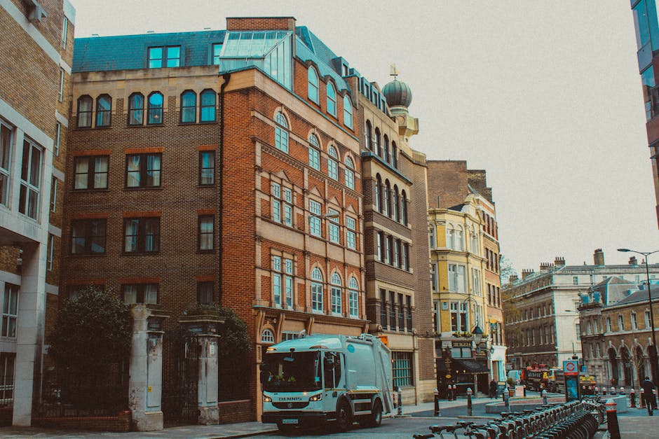 A grey man and van parked on a city street outside a historic brick building with multiple rows of windows, adjacent to other multi-storey buildings with mixed architectural styles, including a yellow-painted facade and decorative elements. The vehicle, positioned near a sidewalk with black bollards and a row of bicycles, is in the process of loading or unloading packing boxes wrapped in brown cardboard and plastic films. Some additional packing materials like bubble wrap and furniture blankets are visible on the area around the van's entrance. The scene is illuminated by natural daylight, suggesting an active home relocation or furniture transport operation, with the presence of [COMPANY_NAME]'s van indicating professional removals services aligned with the guidance on permit requirements for moving in Pimlico.