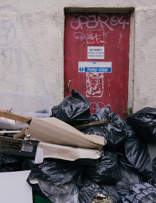 A large pile of black trash bags filled with discarded items, including cardboard boxes and packing materials, is positioned on a pavement in front of a red metal door that has graffiti and multiple signs, such as 'No Money' and 'Keep Clear'. The door is set into a textured, light-colored exterior wall, with some small plants growing at its base. The scene appears to be outdoors in an urban area, possibly near a commercial or residential building, with some natural light illuminating the cluttered environment. In the context of house removals or furniture transport, this scene illustrates the clutter that may be cleared during packing or preparation for a home relocation, with Man and Van Pimlico providing the logistics for moving or clearance services.
