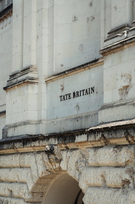 A close-up view of the exterior stone facade of Tate Britain, a historic building with architectural detailing in stone, including decorative molding and weathered surfaces. The photograph shows a section of the building's wall with a prominent sign reading 'TATE BRITAIN' in bold black letters. Below the sign, a weathered stone archway is partially visible, indicating the building's entrance. The scene is captured in natural daylight, highlighting the textures and aged appearance of the stonework. In the foreground, on the pavement outside the building, a man from Man and Van Pimlico is visible engaged in a home relocation process, carefully lifting a large cardboard box wrapped in protective plastic. He is near a small moving van parked adjacent to the pavement, equipped with furniture blankets and straps, ready for furniture transport and packing and moving activities related to house removals in the Pimlico area. The overall setting emphasizes professional moving services supporting efficient home relocation at historic sites like Tate Britain.