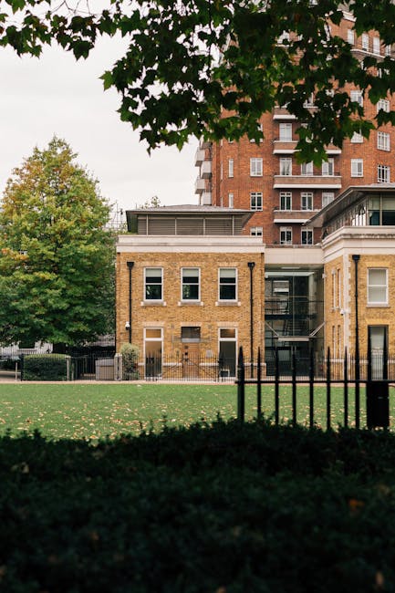A view of a modern, multi-storey residential building with a brick facade and several white-framed windows, situated behind a black metal fence. In the foreground, there is a grassy area with trees and shrubs, some of which have green and yellow foliage, indicating early autumn. The image appears to be taken during daylight, with natural lighting illuminating the scene. This setting could be part of a house relocation process or a move-in/out site managed by Man and Van Pimlico, as part of their removal and moving services in the Tate Britain area, with the building and surroundings prepared for furniture transport and packing activities.